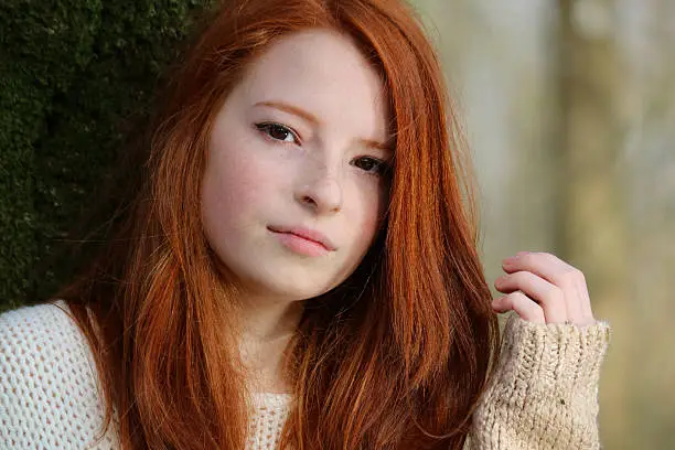 Photo showing a portrait of an attractive, teenage girl with long, flowing, red hair. This picture shows the teenager leaning against the trunk of a tree in a deciduous wood.