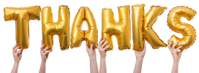 The word thanks has been created by female hands holding individual letter balloons. Each balloon is being held against a plain white background by a caucasian female hand. The Balloons are inflated and made from a shiny reflective gold material.