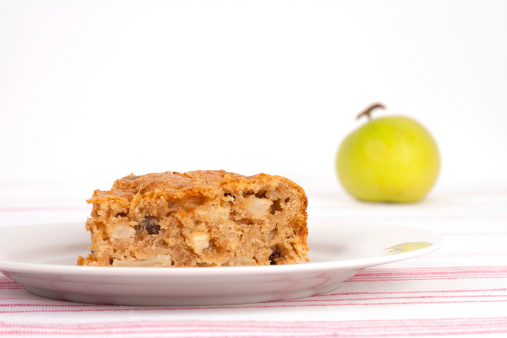 Image of a slice of homemade Dorset apple cake, also containing sultanas, on a white plate, with apples behind.