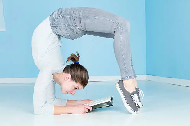 A female gymnast, fit and flexible, reading a book whilst performing a yogic pose