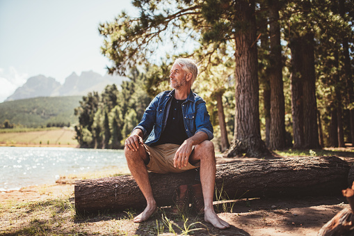Portrait of senior man sitting on a wooden log and looking at the lake. Mature man camping by a lake.
