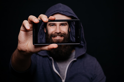 Hipster man with a beard taking picture smartphone self-portrait, screen view, snapshot studio on a black background