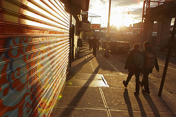 Sunrise down Delancey Street on a New York City morning New York, NY, USA - December 4, 2015: On a typical winter morning as NYC wakes up, two students walk to school east down Delancey Street into the rising sun on the Lower East Side of Manhattan. Steel security shutters are down in front of the as yet unopened local shops. In the distance, commuter cars stream over the Williamsburg Bridge. A sign for an ATM hangs outside a local bodega store. williamsburg bridge photos stock pictures, royalty-free photos & images