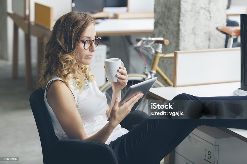 Businesswoman on coffee break in office. Young businesswoman drinking coffee and reading news from digital tablet with feet up. E-Reader Stock Photo