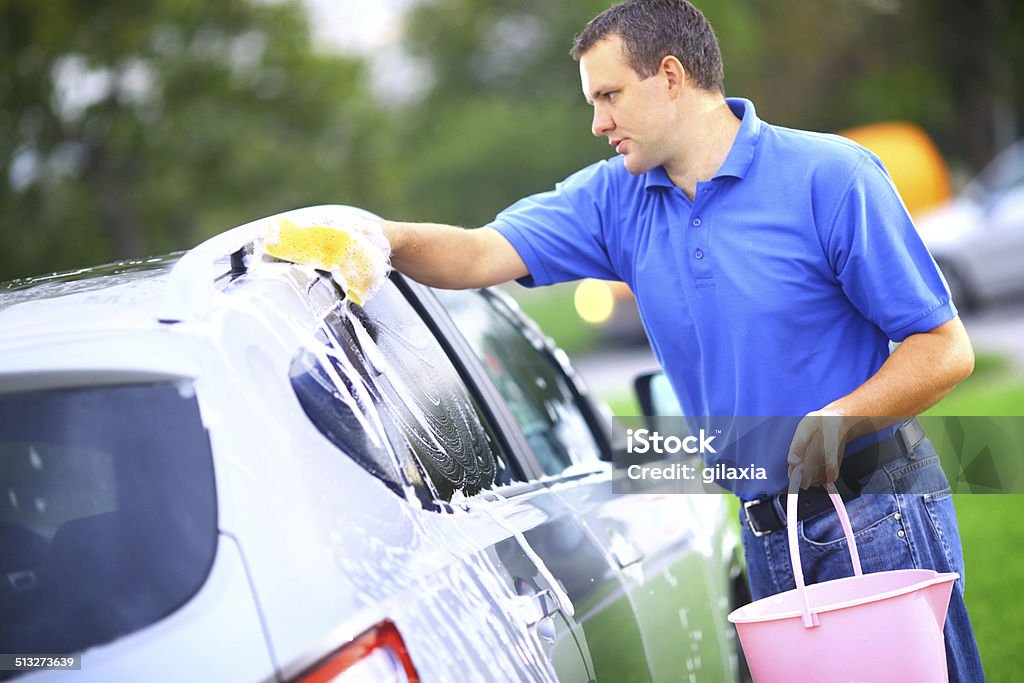 Car wash. Mid-aged man washing a car at parking lot.He's using yellow sponge and bucket with soap.Standing next to his car and washing car roof.Wearing blue polo shirt. Car Stock Photo