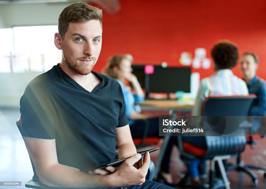 Confident male designer working on a digital tablet in red Casual portrait of a business man using technology in a bright and sunny startup with the team in the background Serious Stock Photo