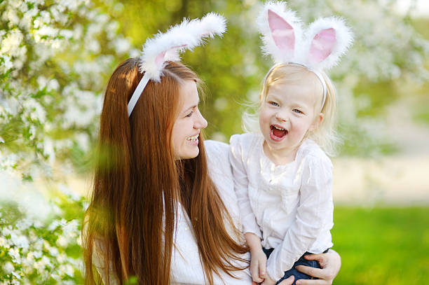 Mother and daughter wearing bunny ears on Easter Young mother and her daughter wearing bunny ears in a spring garden on Easter day holding child flower april stock pictures, royalty-free photos & images
