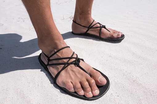 Closeup man's feet wearing primitive black sandals with string laces on white sand beach