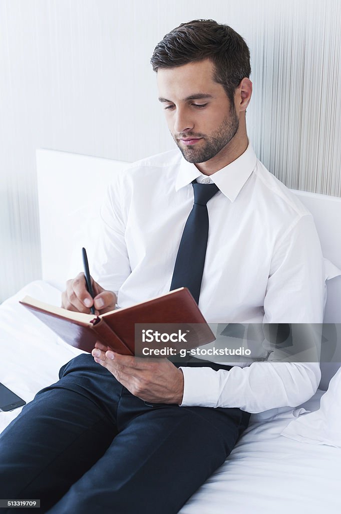 Making some urgent notes. Confident young man in shirt and tie writing something in note pad while sitting in bed at the hotel room Adult Stock Photo