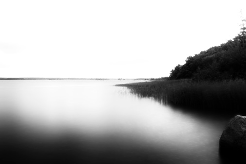 Black / white landscape photograph of a lake. The view is from the water and extends over the entire lake. On the right bank of reeds and trees can be seen. On the horizon, the other side can be imagined. Due to the long exposure the water of the lake appears velvety smooth. 