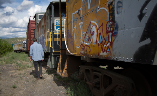 Abandoned graffiti-covered train cars on a track, with man walking alongside. Shot in the Southwest.