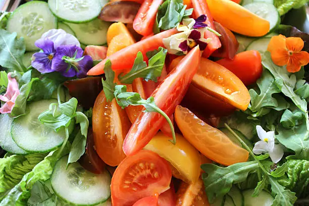 Photo showing a large bowl of green salad, made with lettuce leaves, rocket leaves, slices of cucumber and chunks of homegrown red and yellow tomatoes.  The dish is being served as part of a party food buffet and is pictured garnished with edible flowers (yellow and purple pansies and nasturtiums).