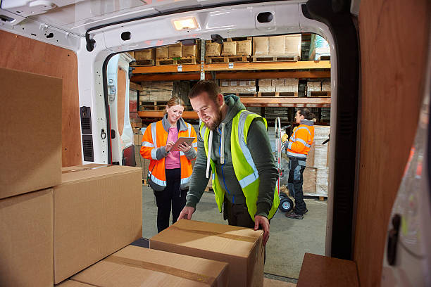 delivery driver in the warehouse a male courier loads or unloads his van supervised by his female manager .. In the background a co-worker can be seen  along the racking. pallet industrial equipment stock pictures, royalty-free photos & images