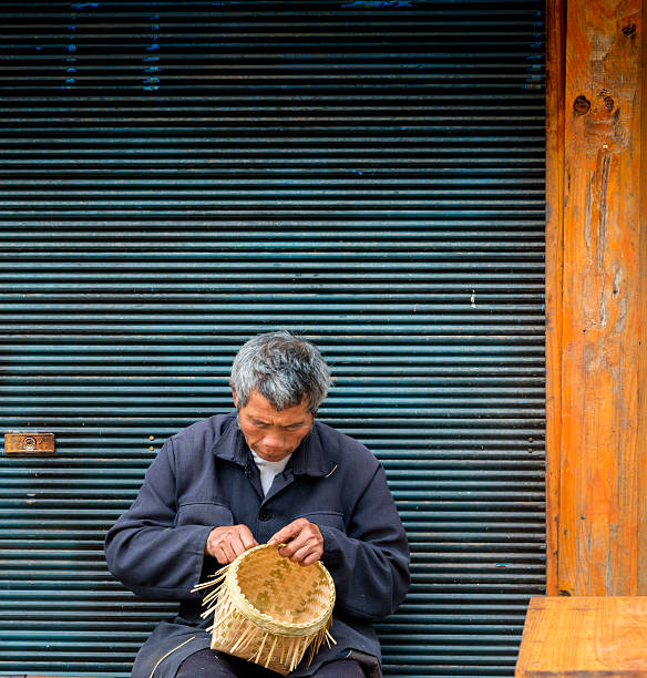 Old man Basket weaving from bamboo, village Zhao Xing. Early morning, Main street of  village Zhao Xing, men is sitting on chair and making a bamboo basket to sell. Used for fruit and other household goods dong stock pictures, royalty-free photos & images