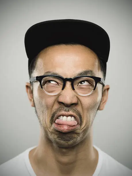 Studio portrait of a japanese young man with bizarre expression. The man is around 30 years and has short hair and casual clothes, wearing a baseball cap and glasses. Vertical color image from a medium format digital camera. Sharp focus on eyes.