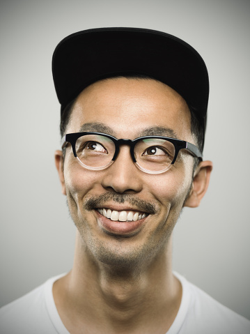 Studio portrait of a japanese young man looking up and very big smile. The man is around 30 years and has short hair and casual clothes, wearing a baseball cap and glasses. Vertical color image from a medium format digital camera. Sharp focus on eyes.
