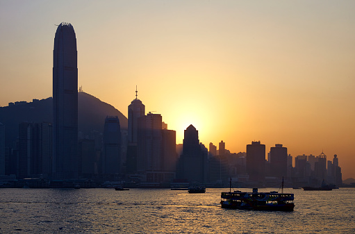 One of Hong Kong's iconic ferries passes the terminal at Tsim Sha Tsui, on its way across the busy waters of Victoria Harbour. In the distance, silhouetted against the setting sun, are the towering skyscrapers of Hong Kong Island.