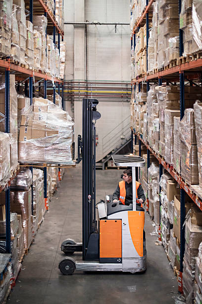 Warehouse worker in uniform loading cardboard boxes with forklift in warehouse full of shelves with goods.