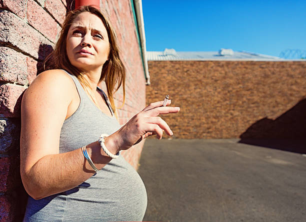 Very pregnant woman sneaks a guilty cigarette A pregnant woman stands in a parking lot as she guiltily sneaks a cigarette, knowing it's bad for her and her baby. Copy space on the tarmac. smoking issues photos stock pictures, royalty-free photos & images