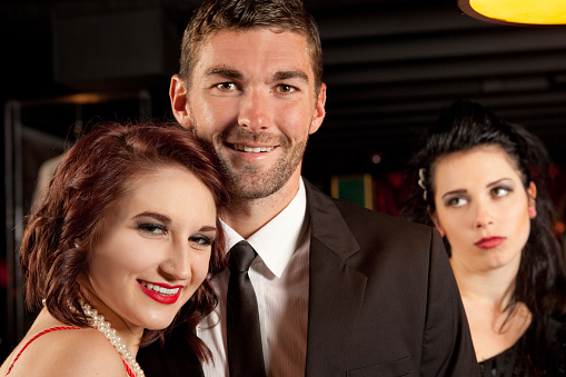 A jealous woman stares at the back of the head of a man and his girlfriend posing for a photo together.