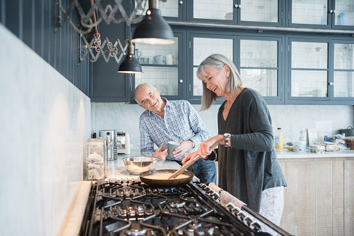 Senior couple making a meal at home in the kitchen. The woman is using a frying pan on the hob talking to her husband, who is smiling and leaning on the kitchen counter