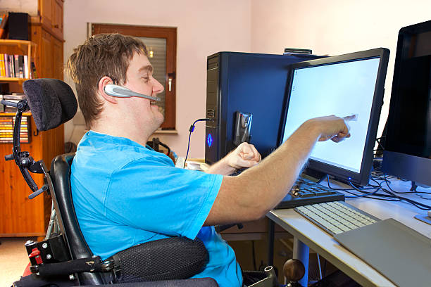 Man with infantile cerebral palsy using a computer. Spastic young man with infantile cerebral palsy caused by a complicated birth sitting in a multifunctional wheelchair, using a computer with a wireless headset, reaching out to touch the touch screen. paraplegic stock pictures, royalty-free photos & images