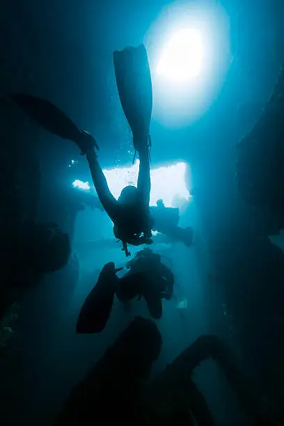 An underwater DSLR photo showing two divers swimming inside the compartments of a sunken ship in Angra dos Reis, Rio de Janeiro, Brazil. The ship is called Pinguino and it sunk in 1967 after a fire. The divers are silhouetted against the blue water and the light coming from above.