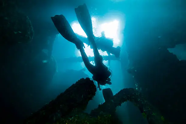 An underwater DSLR photo showing a diver swimming inside the compartments of a sunken ship in Angra dos Reis, Rio de Janeiro, Brazil. The ship is called Pinguino and it sunk in 1967 after a fire. The diver is silhouetted against the blue water and the light coming from above.