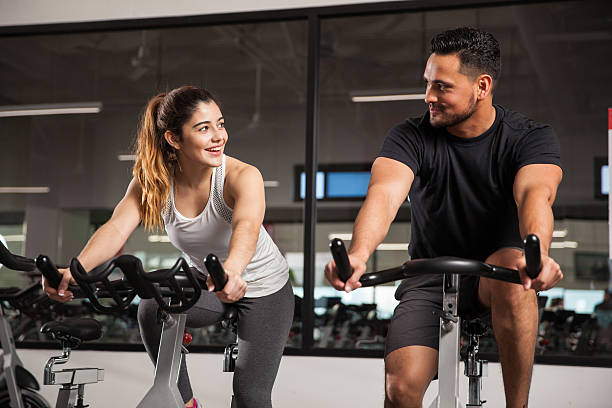 Cute young couple flirting at a gym Beautiful young Hispanic woman flirting and talking to a guy while they both do some exercising at a gym spin bike stock pictures, royalty-free photos & images