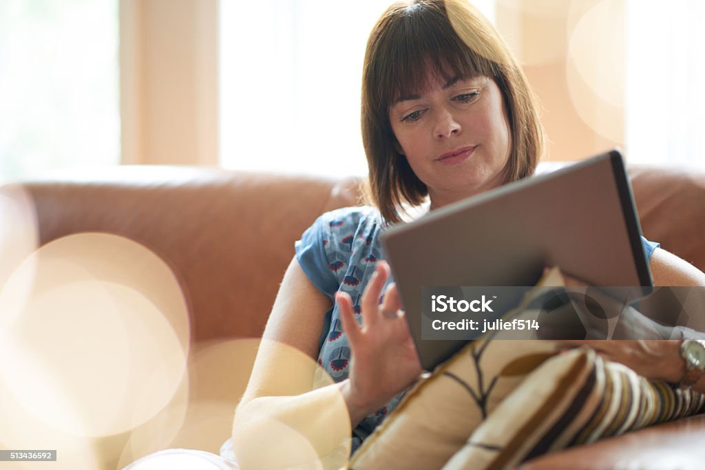 Mature woman browsing the internet on a digital tablet Serene lady in her forties reading emails on a wireless tablet in a brightly lit house- filtered image Digital Tablet Stock Photo