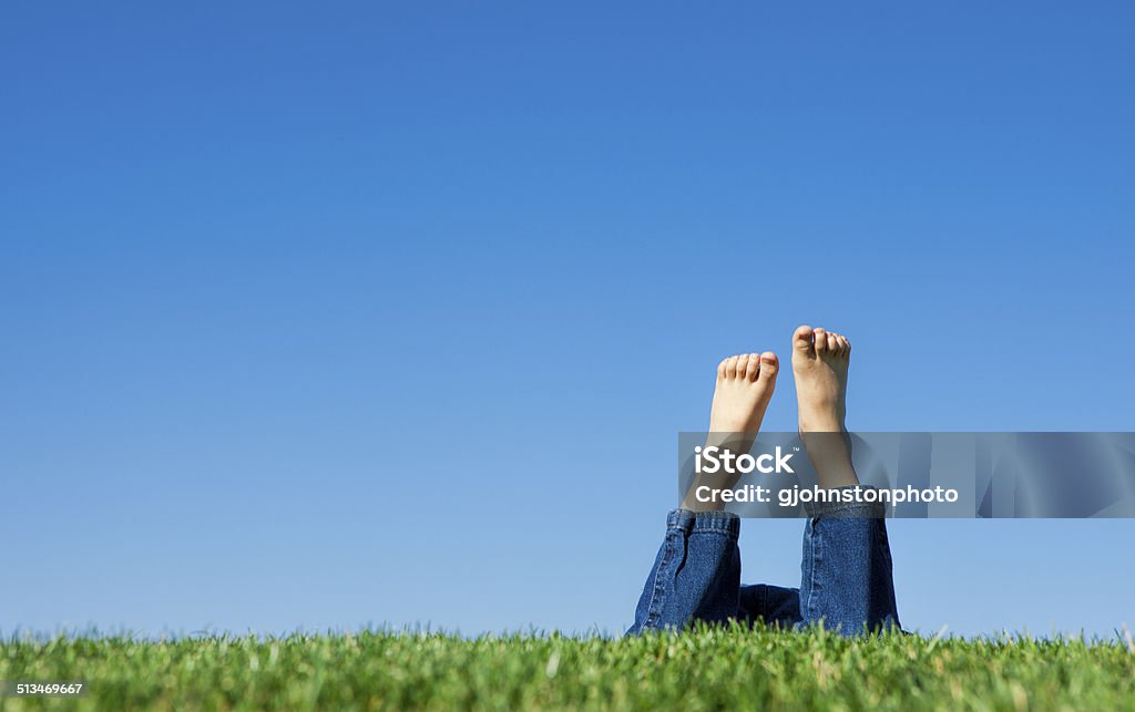 Leisurely laying in the grass. A conceptual image of the feet of a child in the grass shown on a bright summer day against a blue sky. Blue Stock Photo
