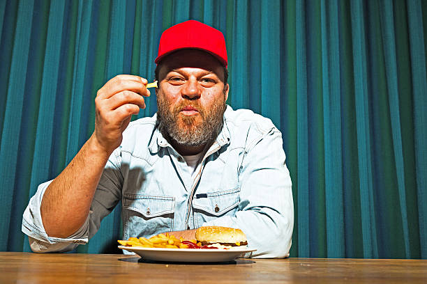 Man with beard eating fast food meal. Enjoying french fries and a hamburger. Trucker with red cap.