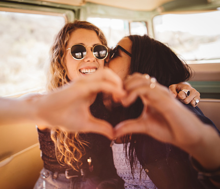 Mixed race hipster girl kisses smiling caucasian friend while both women making a hand heart together for a photo, inside of traveling retro van on road trip.