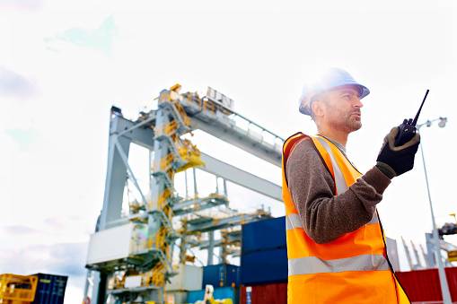 Shot of a young man in workwear talking on a walkie talkie while standing on a large commercial dock