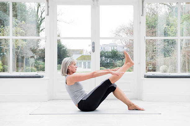 Woman in her 60s stretching leg at home Senior woman sitting on floor with one leg raised and stretching. She is in the conservatory practicing yoga and pilates, keeping fit and exercising. stretched leg stock pictures, royalty-free photos & images