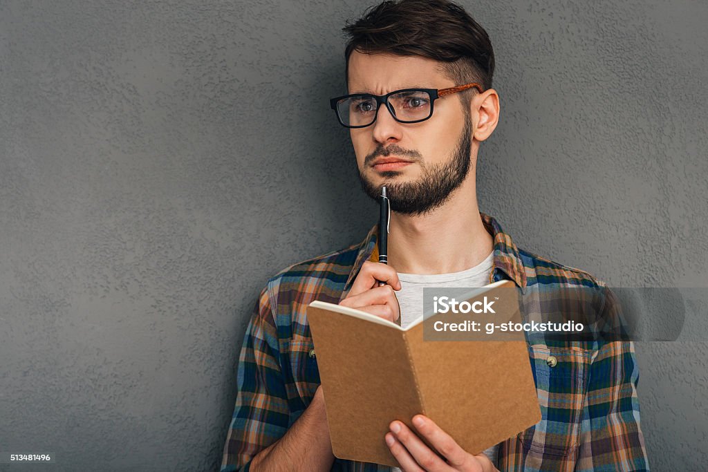 What shall I write? Pensive young man in sunglasses holding notebook and looking away while standing against grey background 20-29 Years Stock Photo