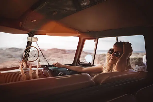 Blonde boho Hipster girl sitting and smiling in front drivers seat of retro van parked at sandy beach during sunset, resting from Road Trip