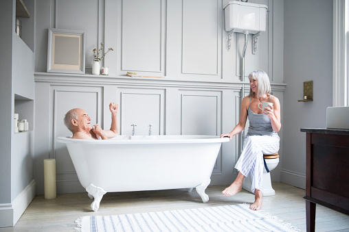 Senior couple in hotel bathroom. Man is taking a bath and woman is sitting with a coffee. They are having a relaxed conversation and are smiling and happy.