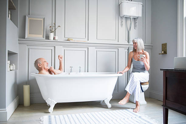 Senior man in bath talking to wife in hotel bathroom Senior couple in hotel bathroom. Man is taking a bath and woman is sitting with a coffee. They are having a relaxed conversation and are smiling and happy. free standing bath photos stock pictures, royalty-free photos & images