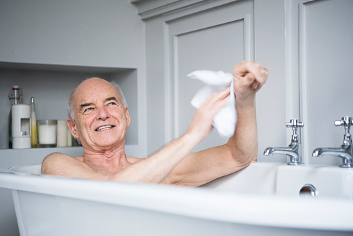 Man in his 70s using a wash cloth to clean his arm in the bath. He is smiling and relaxing in the bath tub, enjoying himself.