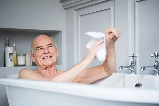 Man in his 70s using a wash cloth to clean his arm in the bath. He is smiling and relaxing in the bath tub, enjoying himself.