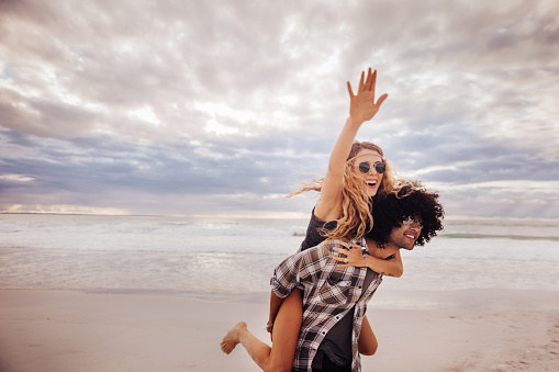 Laughing blonde boho girl piggyback rides on the back of a mixed race hipster man on beach