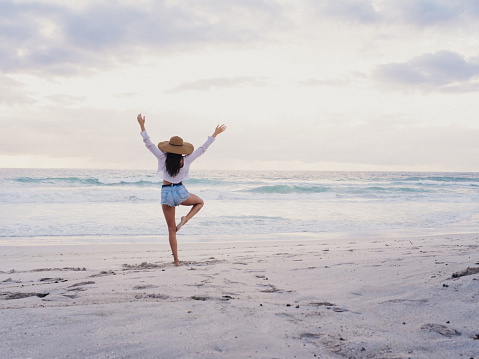 Brunette hipster girl in shorts and hat, practices yoga tree pose while facing the ocean on a sandy beach