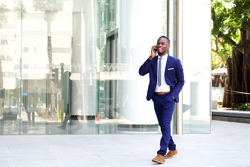 Full length portrait of a happy young businessman walking in the city and talking on mobile phone