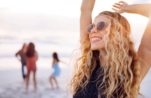 Blonde boho girl raises arms up and smiles enthusiastically in the middle of hipster multi-ethnic group dancing together and moving in front of the ocean on a sandy beach during sunset
