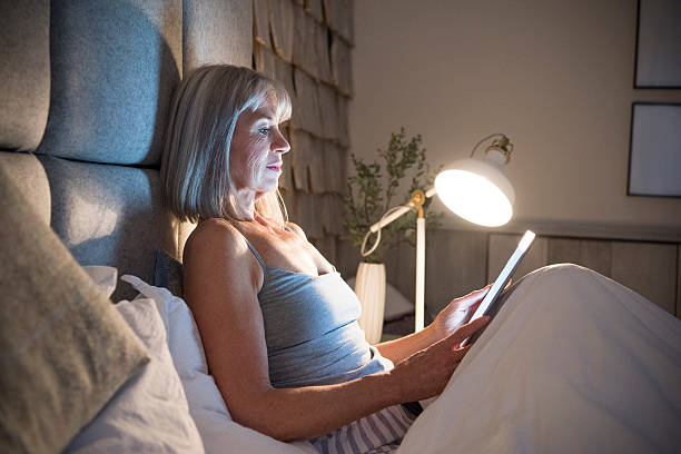 Senior woman sitting in bed at night with tablet Woman in her 60s using digital tablet in bed with electric lamp shining. She is sitting propped up against the headboard reading an ebook. turning on lamp stock pictures, royalty-free photos & images