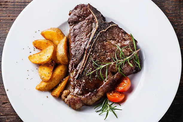 Grilled T-Bone Steak with roasted potato wedges on white plate on wooden background close up