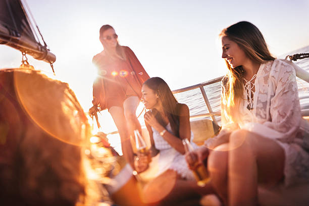 Girl friends drinking beer on sunset yacht cruise with sunflare Group of girl friends happy and relaxing on a golden summer evening, enjoying a sunset cruise on a yacht while drinking beer with brilliant sunflare drinks on the deck stock pictures, royalty-free photos & images