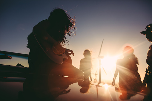 Group of young adult friends is playing guitar and singing during american road trip leaned on their vintage convertible car on a summer sunset under a large wind turbine
