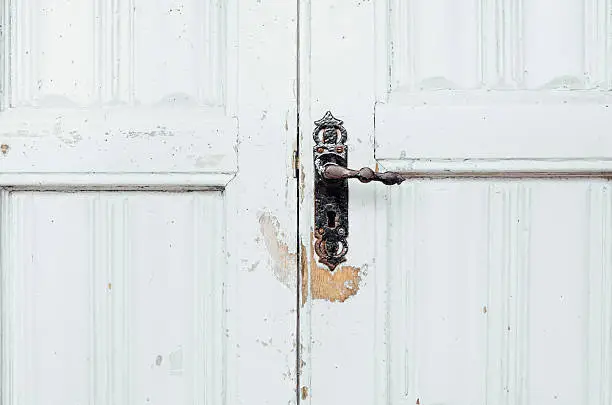 Close-up of a closed old-fashioned wooden double-door with a rusty, black metal door handle with mis-matched screws. The grey paint is chipped and weathered, with an area of bare wood under the keylock, the paint completely worn away by dangling keys. Excellent detail.